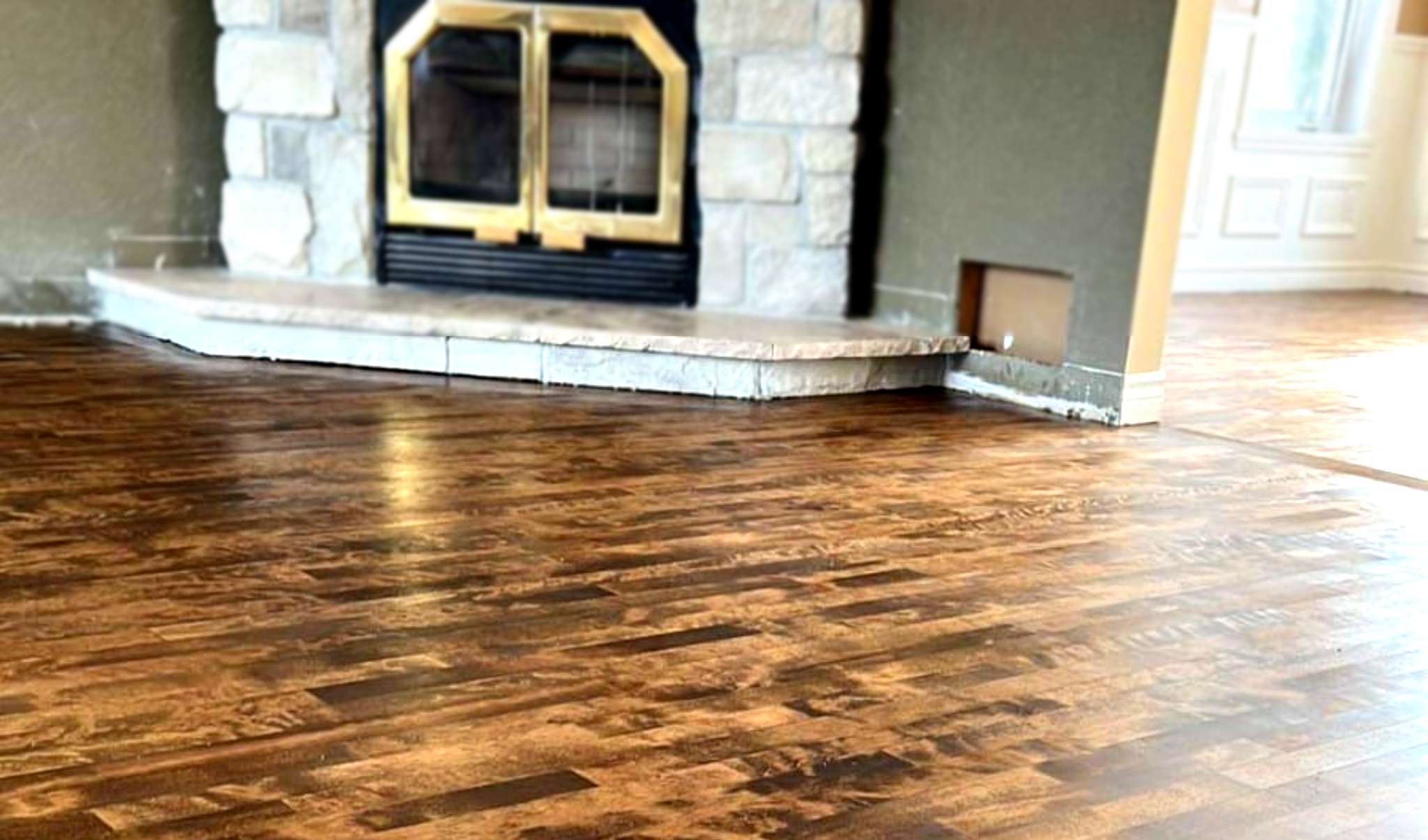 Plancher en bois brun fraîchement rénové dans un salon lumineux, avec une cheminée en pierre et des portes vitrées en arrière-plan, mettant en valeur la texture et la finition brillante du sol.