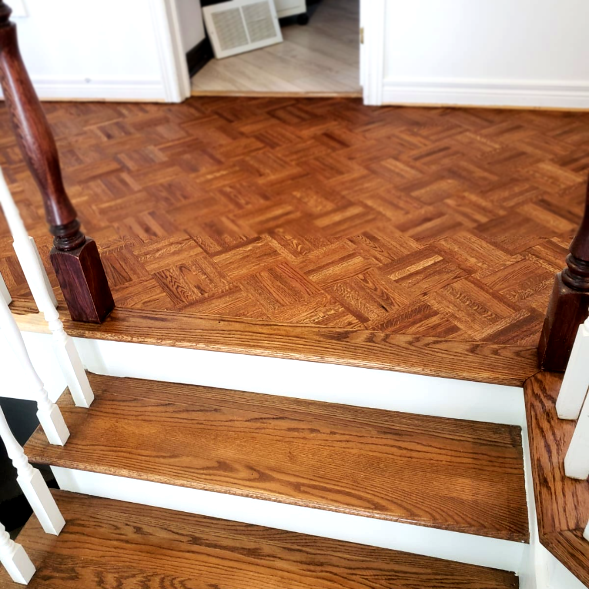 Plancher en bois parquet rénové avec motif géométrique, situé sur un palier d’escalier avec marches en bois et murs blancs, mettant en valeur une finition brillante et uniforme.
